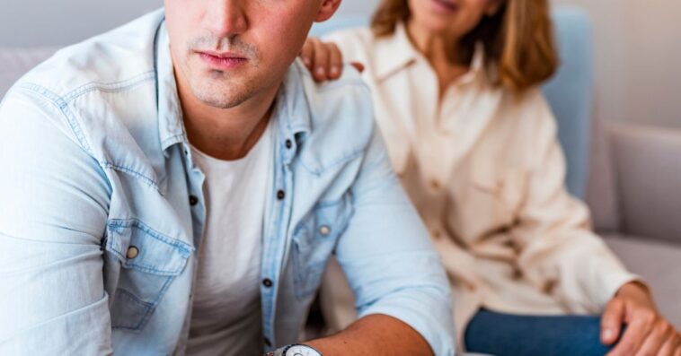 Mature Mother and problem child. Frustrated mother talking to with her upset son sitting on the couch. Senior mother talking with her unhappy son at home during the day. Mother comforting her black hair sad son.