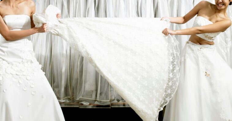 Young women in fitting room tugging at gown.