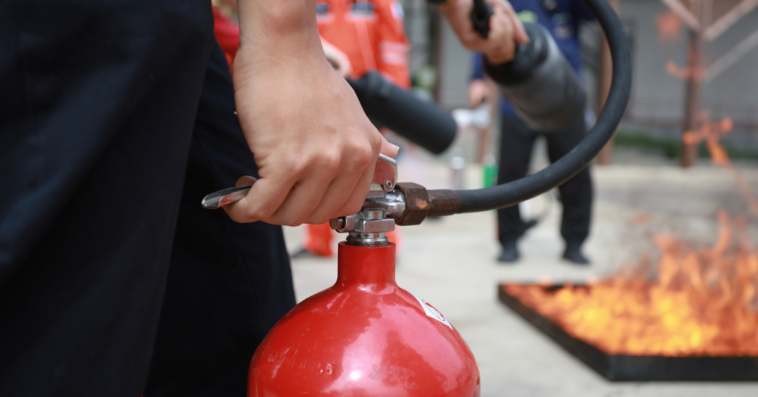 A hand holding a fire extinguisher with a fire in the background.