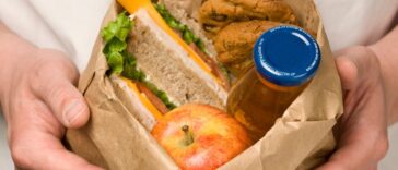 Hands holding a bagged lunch with a sandwich, apple, cookies and juice.
