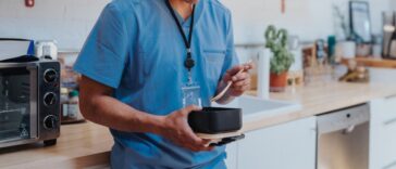 A young adult male nurse leans against the kitchen counter in a hospital break room, eating lunch during a work break.