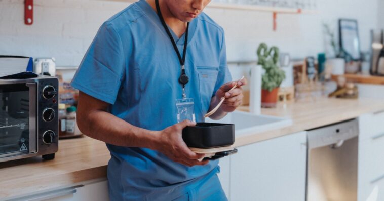 A young adult male nurse leans against the kitchen counter in a hospital break room, eating lunch during a work break.