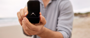 A man holding out an open ring box with a diamond ring inside it.