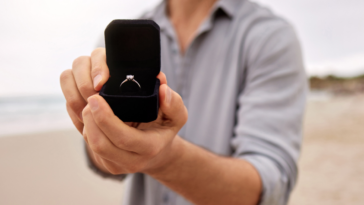 A man holding out an open ring box with a diamond ring inside it.