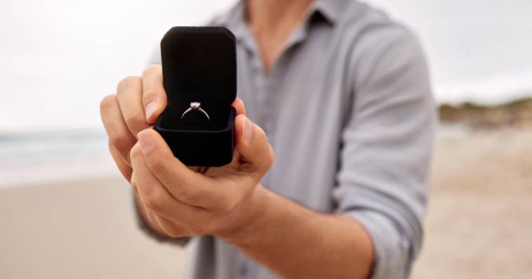 A man holding out an open ring box with a diamond ring inside it.