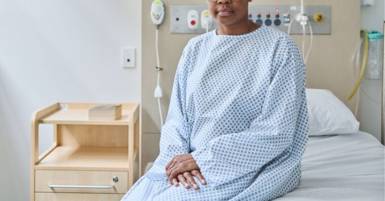 Portrait of a bald woman suffering from cancer sitting on bed at hospital. Female patient is wearing medical gown. She seems to be serious.