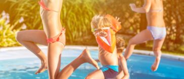 Four kids, one with water wings jumping into a pool in the backyard.