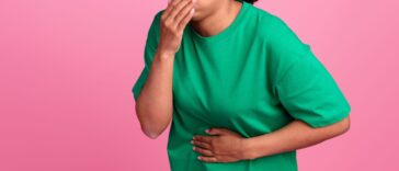Photo portrait of a pretty young girl as she covers her mouth. She suffera nausea and wears a trendy green outfit isolated on a pink color background.