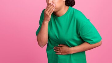 Photo portrait of a pretty young girl as she covers her mouth. She suffera nausea and wears a trendy green outfit isolated on a pink color background.