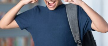 Teenager boy carrying bag in library, has his fingers in his ears.