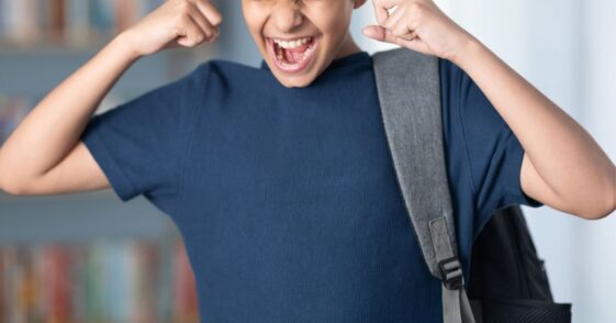 Teenager boy carrying bag in library, has his fingers in his ears.