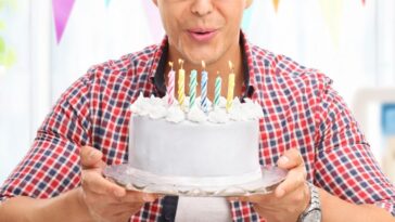 Joyful young man blowing candles on a birthday cake and looking into the camera at home.