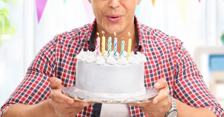 Joyful young man blowing candles on a birthday cake and looking into the camera at home.
