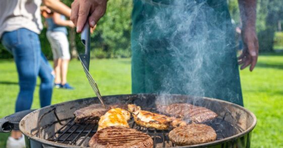 Mid section of man turning burgers and meat on barbecue grill in garden.
