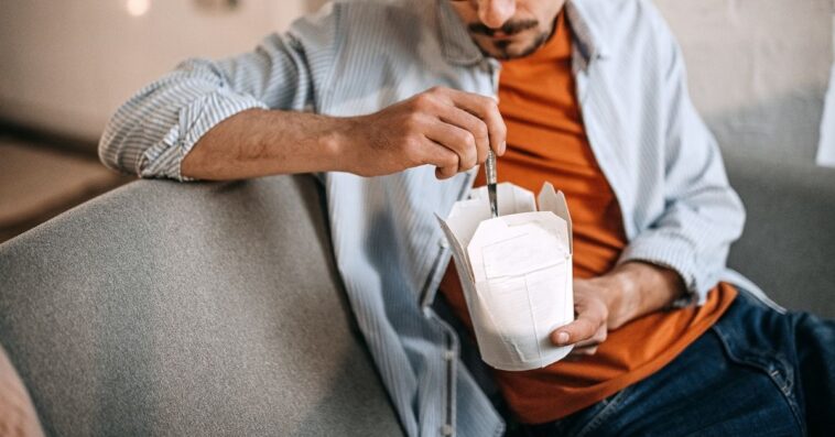 A young man with glasses sits on a couch eating out of a carton.