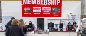 A Costco membership counter where customers are being assisted by employees, with a large sign displaying membership options.