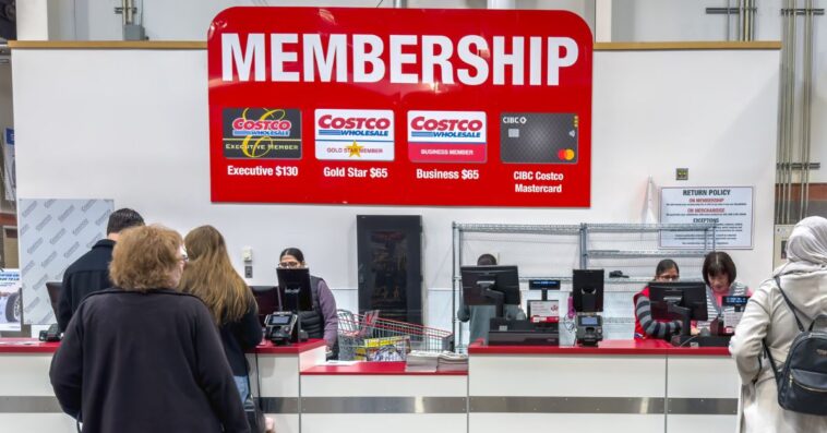 A Costco membership counter where customers are being assisted by employees, with a large sign displaying membership options.