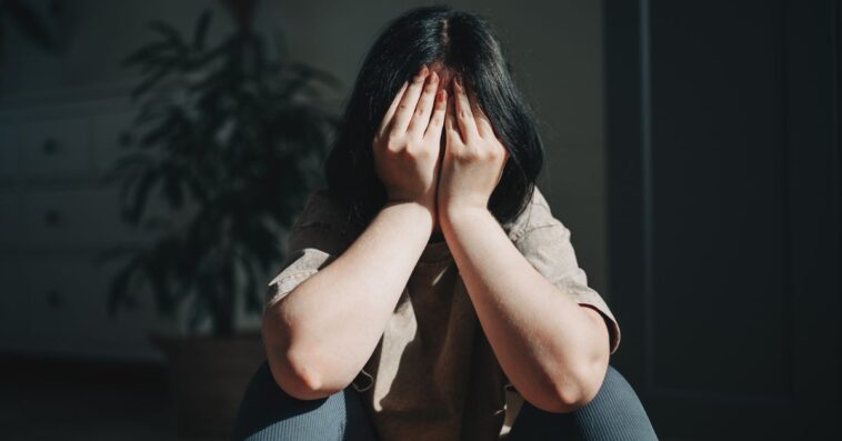 Upset teen girl sits on the floor, covering her face with her hands.