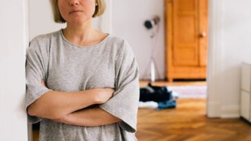 Serious portrait of woman standing against a wall in her home, her arms are crossed.