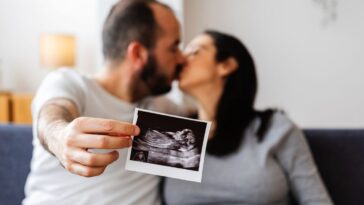 Young, happy couple sharing a loving kiss on the sofa, proudly holding and displaying their ultrasound scan of a baby on the way.