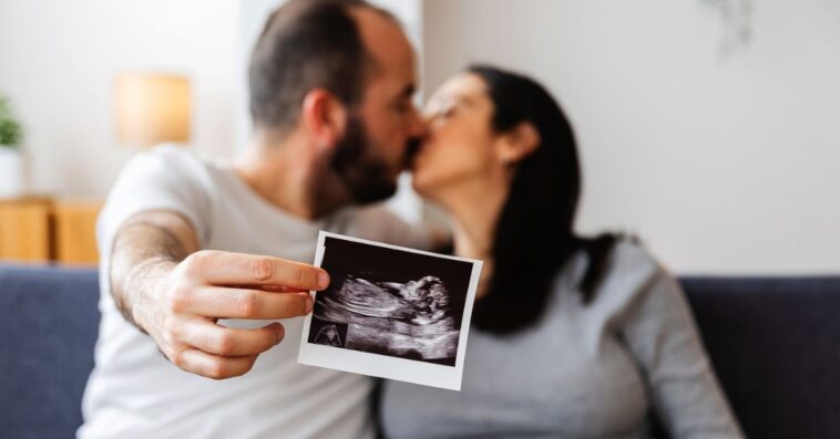 Young, happy couple sharing a loving kiss on the sofa, proudly holding and displaying their ultrasound scan of a baby on the way.