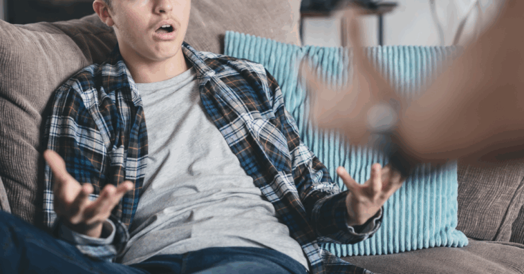 A teenage boy sitting on the couch holding out his arms and hands.