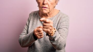 A pretty female senior citizen angrily looks into the camera and points. She stands in front of an isolated pink background.