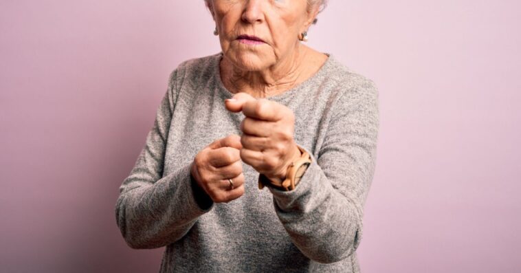 A pretty female senior citizen angrily looks into the camera and points. She stands in front of an isolated pink background.