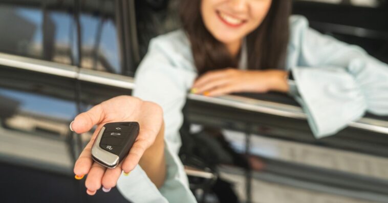 A woman's hand holds out a key to new car, through the open passenger side window.
