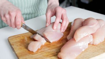 Unidentifiable hands cut up raw chicken breasts on a cutting board.