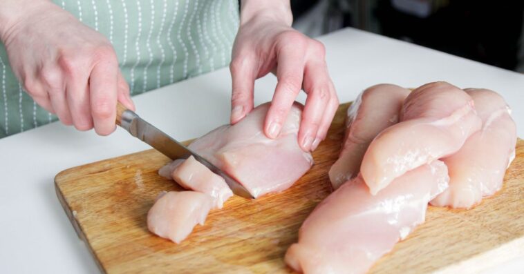 Unidentifiable hands cut up raw chicken breasts on a cutting board.