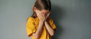 A sad, young girl cries into her hands. She wears a yellow shirt and stands in front of a gray background.