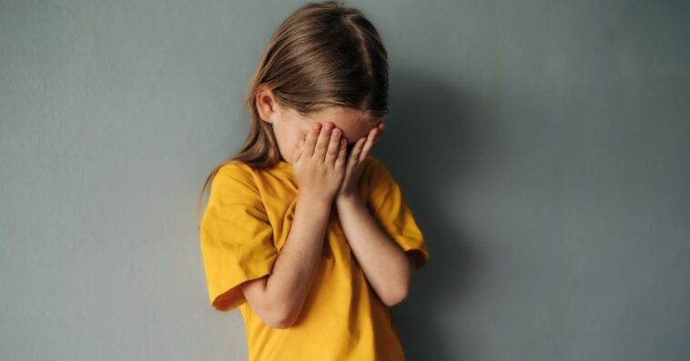 A sad, young girl cries into her hands. She wears a yellow shirt and stands in front of a gray background.