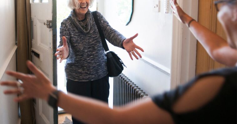 Mature lady entering front door of house, holding out her arms to hug another woman.