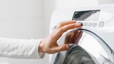A cropped shot of a woman's hand turning on an automatic washing machine or select program with a button on the control panel in a white bathroom in a modern house.