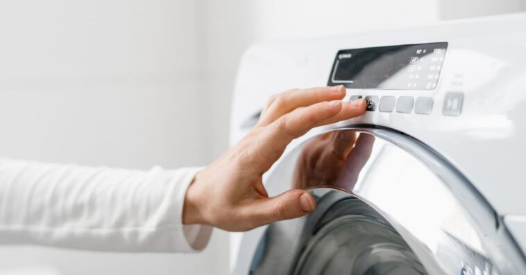 A cropped shot of a woman's hand turning on an automatic washing machine or select program with a button on the control panel in a white bathroom in a modern house.