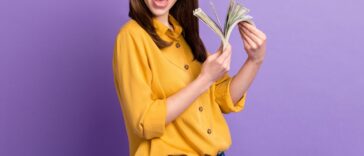 A young, smiling woman fans dollar bills while standing in front of a purple background.