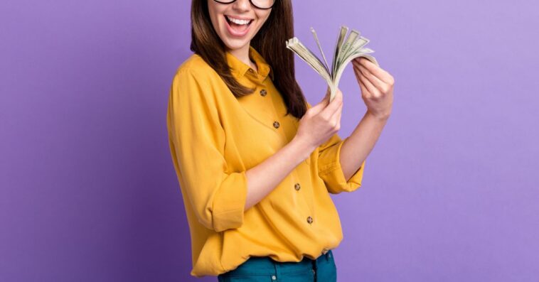 A young, smiling woman fans dollar bills while standing in front of a purple background.