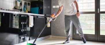 A man cleaning the floor in a modern kitchen and living area using a cordless vacuum cleaner. The room is bright with natural light, featuring a contemporary design with reflective black cabinets, a wall calendar, and large windows. The man is casually dressed and focused on cleaning, with the vacuum's green laser light illuminating the floor.