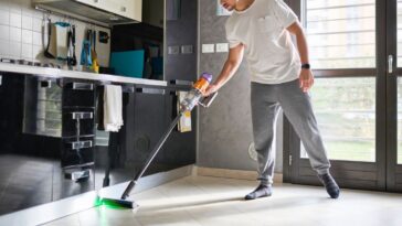 A man cleaning the floor in a modern kitchen and living area using a cordless vacuum cleaner. The room is bright with natural light, featuring a contemporary design with reflective black cabinets, a wall calendar, and large windows. The man is casually dressed and focused on cleaning, with the vacuum's green laser light illuminating the floor.