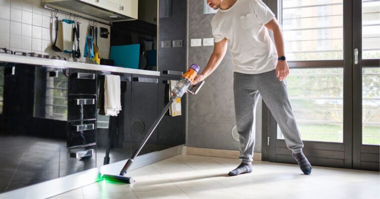 A man cleaning the floor in a modern kitchen and living area using a cordless vacuum cleaner. The room is bright with natural light, featuring a contemporary design with reflective black cabinets, a wall calendar, and large windows. The man is casually dressed and focused on cleaning, with the vacuum's green laser light illuminating the floor.