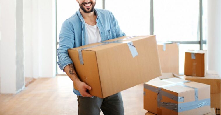 Closeup front view of a smiling young man carrying boxes as he moves into his new apartment.