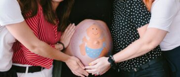 Group of smiling, female friends surrounding a pregnant woman at a baby shower. There is a baby drawn on the mother's belly.