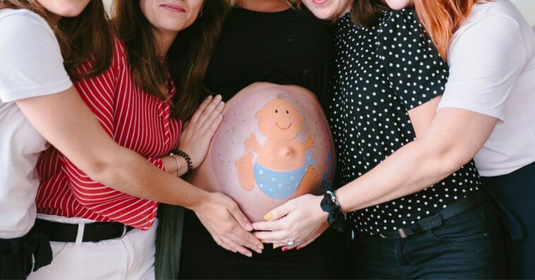 Group of smiling, female friends surrounding a pregnant woman at a baby shower. There is a baby drawn on the mother's belly.