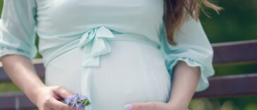 Close up of beautiful young woman, sitting on a bench during the day. She is in her last trimester of pregnancy, wearing a mint color, vaporous summer dress and holding flowers in her hands in a caressing gesture around her baby bump.