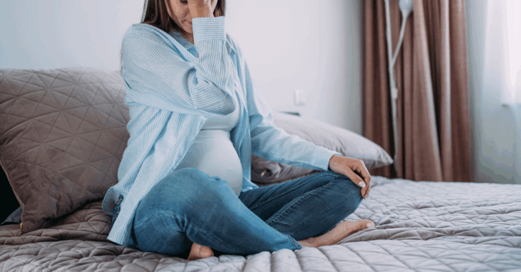 A pregnant woman sitting on a bed, resting her head in her hands.