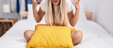 A young blonde woman sitting on the bed, with a pillow, at home, crazy and mad, shouting and yelling with aggressive expression and arms raised.