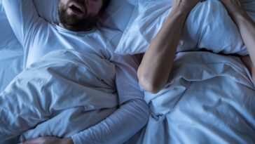 A stressed woman, covers her face with a pillow, she cannot fall asleep because her husband is snoring in bed at night.