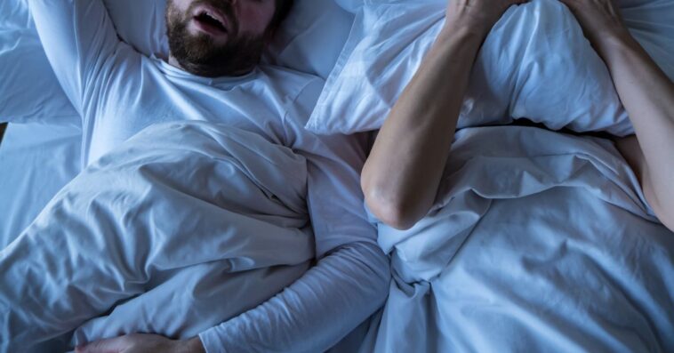 A stressed woman, covers her face with a pillow, she cannot fall asleep because her husband is snoring in bed at night.