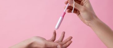 A close up on a pink background with two hands of women passing a tampon in applicator.